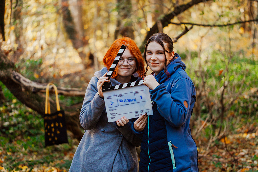 Zwei junge Mädchen stehen neben einander in einem wald. Sie halten gemeinsam eine Filmklappe in den Händen. 
